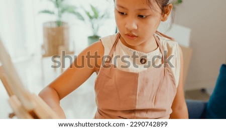 Close-up Asian toddler girl with apron sit on sofa chair in front of painting canvas with color palette use paint brush focus on painting abstract on weekend at home. Creative lifestyle kid concept.