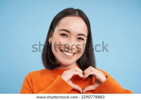 Close up portrait of lovely asian woman, shows heart sign and smiles with care and tenderness, stands over blue background.