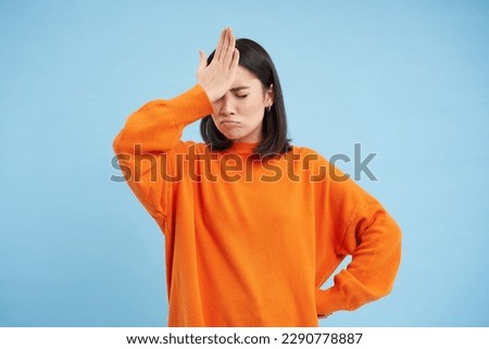 Epic fail. Disappointed asian woman slaps her forehead with upset face, forgot smth, annoyed, stands over blue background in orange sweatshirt.