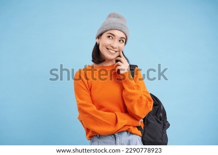 Carefree beautiful woman in streetwear, holds backpack, smiles and looks thoughtful, blue background.