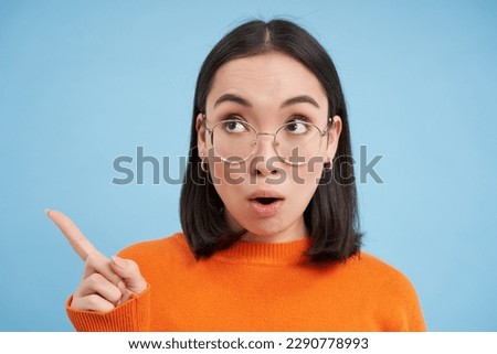 Close up portrait of woman in glasses, points and looks left, shows advertisement, stares at banner with surprised face, blue background.