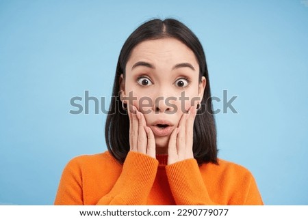 Close up portrait of amazed brunette girl says WOW, amazed by smth. Asian woman looks surpirsed at camera, impressed reaction, blue background.
