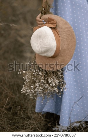 Girl in long light blue dress holding beige hat and bouquet of daisies