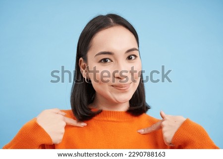 Close up of confident smiling korean girl, points at herself with pleased, satisfied face, stands over blue background. Copy space