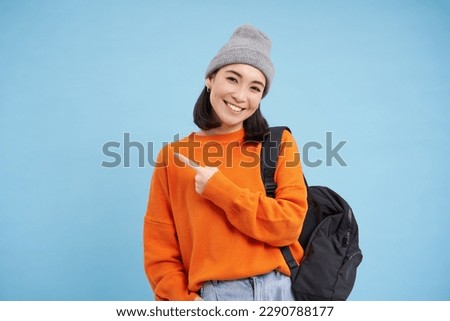 Enthusiastic asian woman, points finger left at banner, shows promo offer, smiles and looks happy, stands in hat with backpack, blue background.