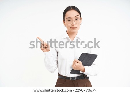 Portrait of korean corporate woman in glasses, holds digital tablet, points finger left and looks strict and serious, stands over white background. Royalty-Free Stock Photo #2290790679
