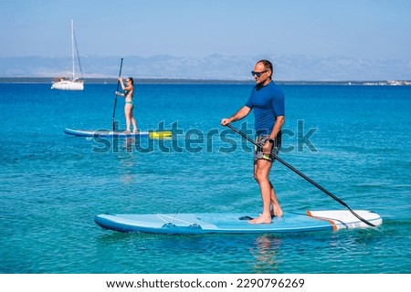 Couple riding SUP stand up paddle on vacation. Active man and woman riding SUP boards and paddling in the ocean on a sunny day. Professional SUP instructor teaching. Taking lessons in Silba, Croatia. Royalty-Free Stock Photo #2290796269
