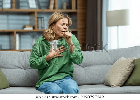 An attack of asthma, allergies at home. An elderly woman is sitting on the sofa, holding her chest and coughing, feeling severe pain, suffocating. Royalty-Free Stock Photo #2290797349