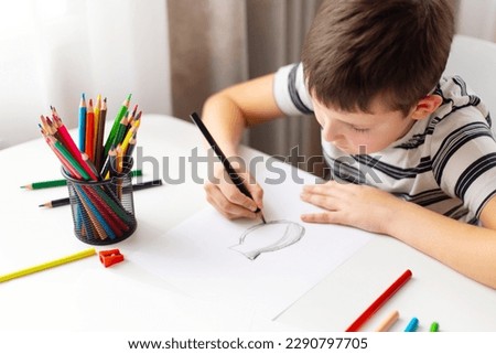 A child boy draws on white paper with colored pencils while sitting at a table