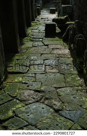 A photo of an ancient stone path leading through the Banteay Kdei Temple in Angkor, Cambodia. Bright green moss grows on the old sandstone blocks which are surrounded by shadows. 