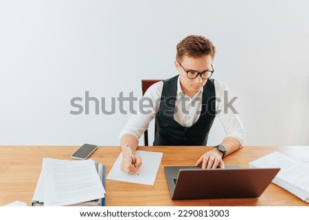 A young white-collar office worker is busy writing on a paper while wearing eyeglasses in his modern home office. The laptop and pen are placed neatly on his desk.