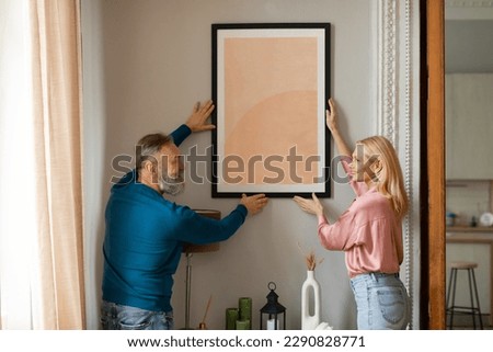 Happy Mature Couple Hanging Picture In Frame On Wall Together Standing Indoors. Spouses Decorating Their Cozy House With Poster. Art And Interior Design Decoration Concept
