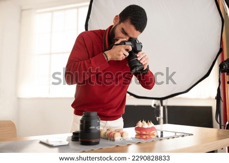 Handsome hispanic guy in casual professional photographer taking photo of tasty dessert cake and macarons, photo studio interior, young man holding digital camera, copy space