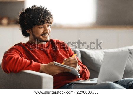 Online Education. Handsome young indian man using laptop and taking notes while sitting on couch at home, smiling eastern guy watching tutorial, lecture or webinar, looking at computer screen Royalty-Free Stock Photo #2290839533