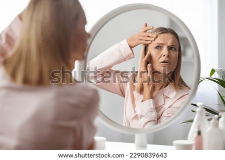 Upset middle aged woman checking wrinkles around her eyes while making beauty routine at home, beautiful mature lady looking in mirror and touching face, feeling tired and stressed, selective focus Royalty-Free Stock Photo #2290839643