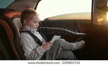 Child girl in playing an online game with digital tablet while sitting in back seat of car during trip. Happy child enjoys vacation with tablet pc in car, family trip. Daughter travels with computer Royalty-Free Stock Photo #2290851741