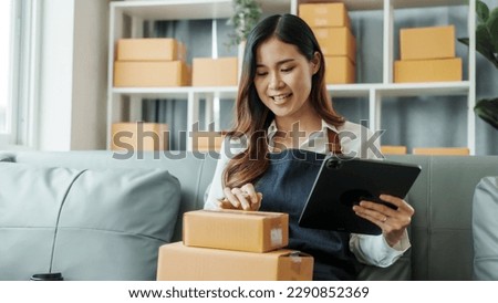 Using tablet computer, Young pretty Asian female influencer small businesses SME owners female entrepreneurs working on receipt box and check online orders, sitting on sofa of home online business.