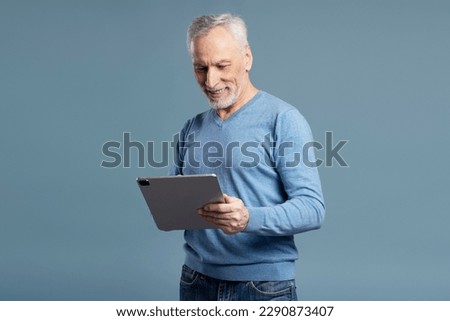 Handsome smiling senior man holding digital tablet shopping online isolated on blue background. Happy 60 years old male watching videos, reading e book. Technology concept  