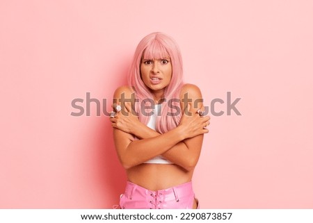 Young woman in pink wig posing isolated against studio wall, dressed in a sleeveless top looks like she is cold, hugging herself, copy space, high quality photo