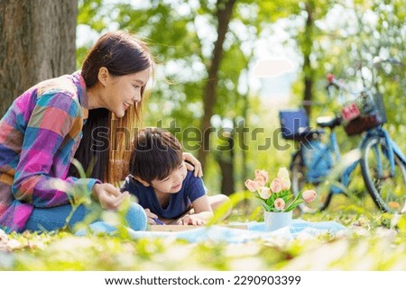 Happy cheerful Asian lovely little boy and mother sitting under the tree at park in the morning and drawing a picture. Asian woman spending time in weekend with her lovely little son.