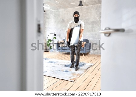 Masked face. Man burglar stealing tv set from house. Thief with black balaclava stealing modern expensive television.  Royalty-Free Stock Photo #2290917313