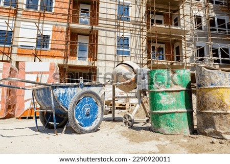 Industrial obsolete wheelbarrow uses for transportation burden and cement mixer machine are at construction site. Building with scaffolding is under construction.