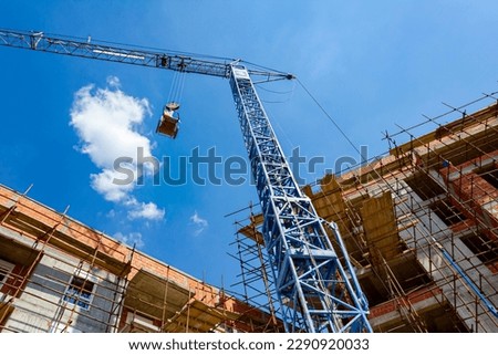 Shot from below of crane that puts down on ground wheelbarrow full of industrial waste, crumpled cellophane, in background is edifice under construction with scaffold and blue clear sky. Royalty-Free Stock Photo #2290920033