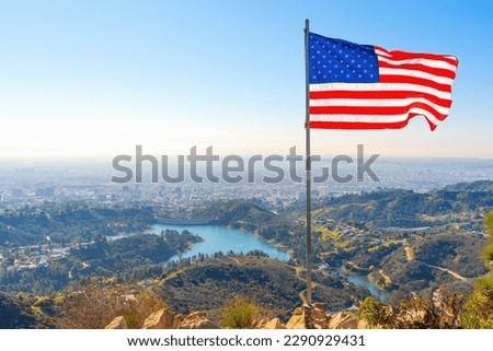 American flag waving in the wind, set against the stunning backdrop of Hollywood Lake, majestic mountains and the bustling cityscape of Los Angeles as seen from the top of the Griffith Park Trail.