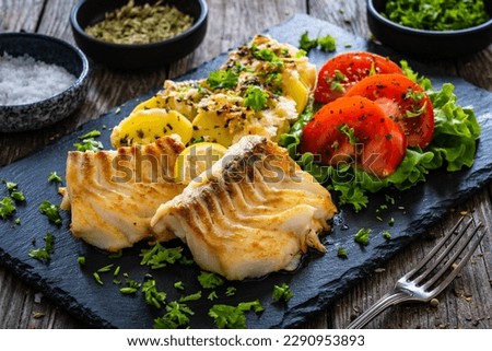 Fried cod loin with baked potatoes and fresh vegetables on black plate on wooden table 