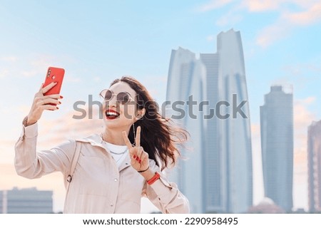 With a confident smile and a smartphone in hand, a girl captures a beautiful moment in front of the towering skyscrapers of Abu Dhabi.