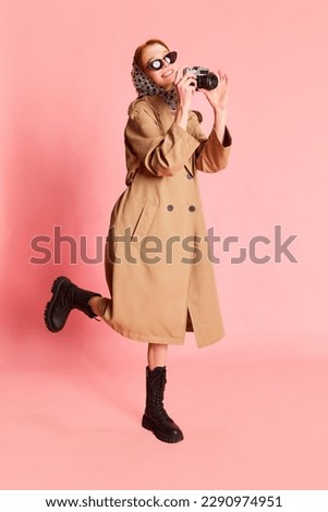 Photo hunter. Portrait of stylish girl, woman wearing stylish elegance look with headscarf, sunglasses, trench coat taking photo and smiling over pink background. Fashion, beauty, emotions concept