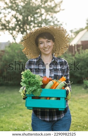 Farmer woman holding wooden box full of fresh raw vegetables. Basket with vegetable cabbage, carrots, cucumbers, radish, salad, garlic and pepper in female hands. Royalty-Free Stock Photo #2290982525