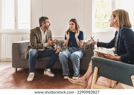 Couple Arguing During Marriage Counseling - Married couple heatedly arguing on a couch during family therapy. The woman looks frustrated, while the man appears aggressive. Psychologist mediates. Royalty-Free Stock Photo #2290983827