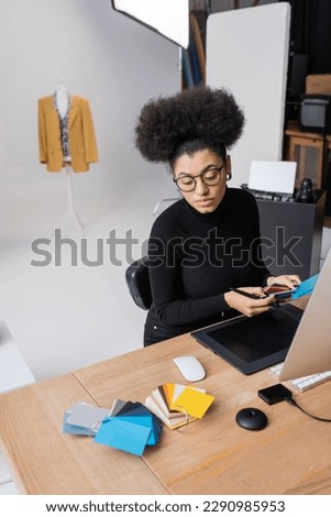 african american retoucher in eyeglasses holding color swatches near graphic tablet and computer monitor in photo studio