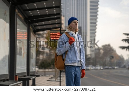 Upset backpacker student stylish guy citizen urbanite waiting bus on bus stop in downtown in spring overcast day. Problem of public transport in city concept. Modern human friendly city environment.  Royalty-Free Stock Photo #2290987521