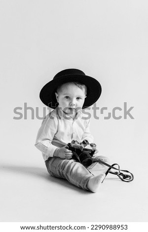 Children's studio portrait. Child in hat sitting on floor and play with photo camera. World Photographers Day. Fun kid holds retro vintage camera isolated on white wall. Mockup. Black and white photo.