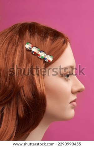 Close-up shot of a young European woman with red hair with a colorful alligator hair clip in her hair. A girl with resin hair clip is posing on a pink background, looking to the side. Side view.      Royalty-Free Stock Photo #2290990455