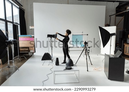 full length of african american content producer in black clothes assembling lighting equipment in spacious photo studio Royalty-Free Stock Photo #2290993279