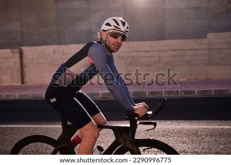Fit male athlete on time trial aero bike in a relaxed position, riding his bike during recovery after doing a hard session of anaerobic training. Man riding triathlon bike in cycling kit and helmet. Royalty-Free Stock Photo #2290996775
