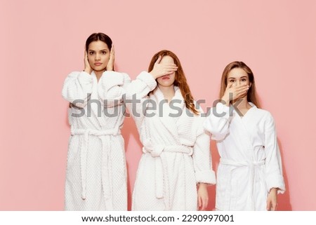 Three girls in bathrobes covering eyes, mouth and ears, posing against pink studio background. Keeping secrets. Concept of youth, face care, beauty, friendship, party, relaxation, sleepover
