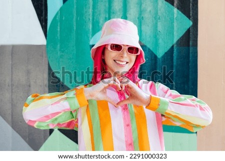 Vanilla Girl. Kawaii vibes. Candy colors design. Young woman with pink hair and sun glasses in bucket hat and multicolor shirt making sign Heart by fingers on the wall background. Love fashion concept Royalty-Free Stock Photo #2291000323
