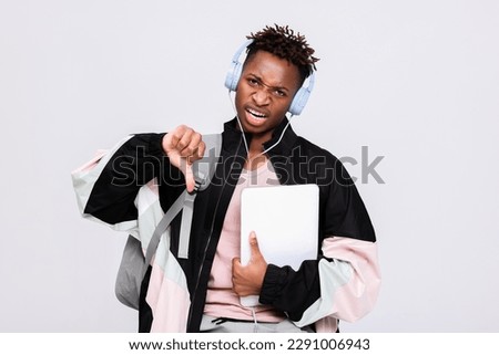 Sad angry dissatisfied afro american dark skinned guy man wearing new modern headphones listening to music ot ready to come back to school university holding backpack rucksack and laptop in hands.