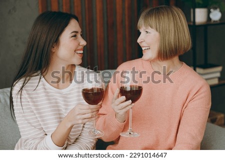 Two smiling happy fun adult women mature mom young kid wearing casual clothes drink wine put sit on gray sofa couch stay at home flat rest relax spend free spare time in living room. Family concept