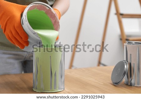 Woman pouring green paint from bucket into can at wooden table indoors, closeup