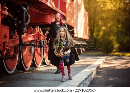A woman accompanies her child to the school of magic. A girl in a raincoat, in a scarf, with a suitcase near the red train. Photo with a locomotive. Halloween images