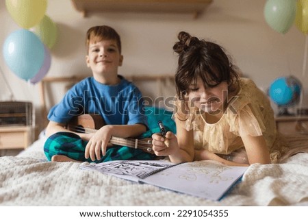 Little boy plaing on the guitar, while his sister drawing a picture.