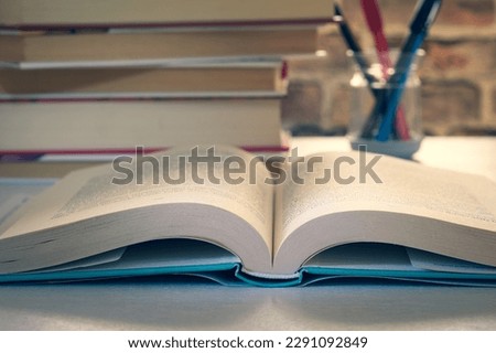 An open hardcover book on a desk, in the background a pile of books and a pen holder, world book day concept. Royalty-Free Stock Photo #2291092849