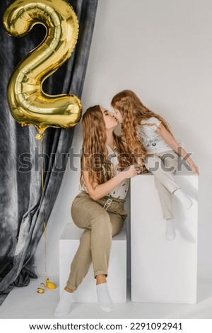Mother congratulates daughter, who has entered second grade at school. 2nd class. Happy schoolgirl near white wall with gel balloon in studio. Mom kisses kid girl. Happy family having fun celebrating. Royalty-Free Stock Photo #2291092941