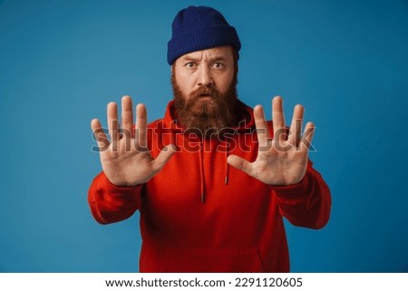 Bearded man wearing blue hat and red hoodie showing stop sign with palms isolated over blue background