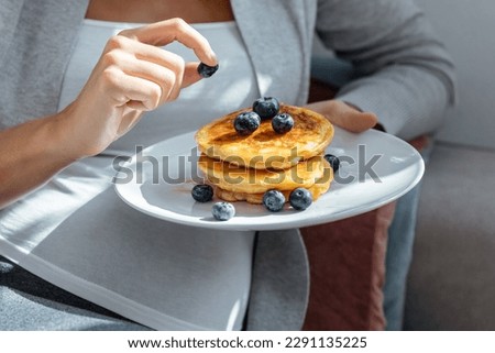 Shot of beautiful happy pregnant woman in panties eating pancakes with red fruits lying on sofa at home